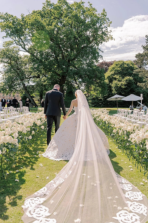 Wedding processional as bride and groom walk hand in hand down a rose petal aisle, her long veil trailing past white chairs on a garden lawn