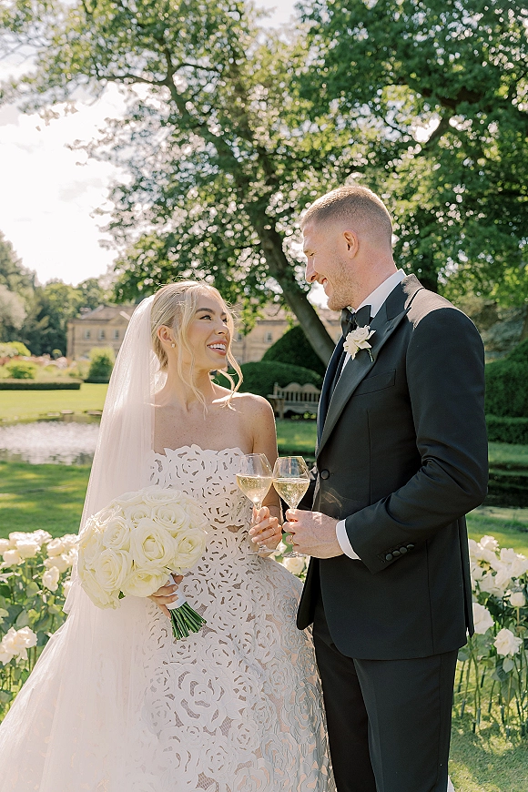 Couple portrait of bride and groom toasting with champagne flutes, bride holding a white rose bouquet in a garden by a pond