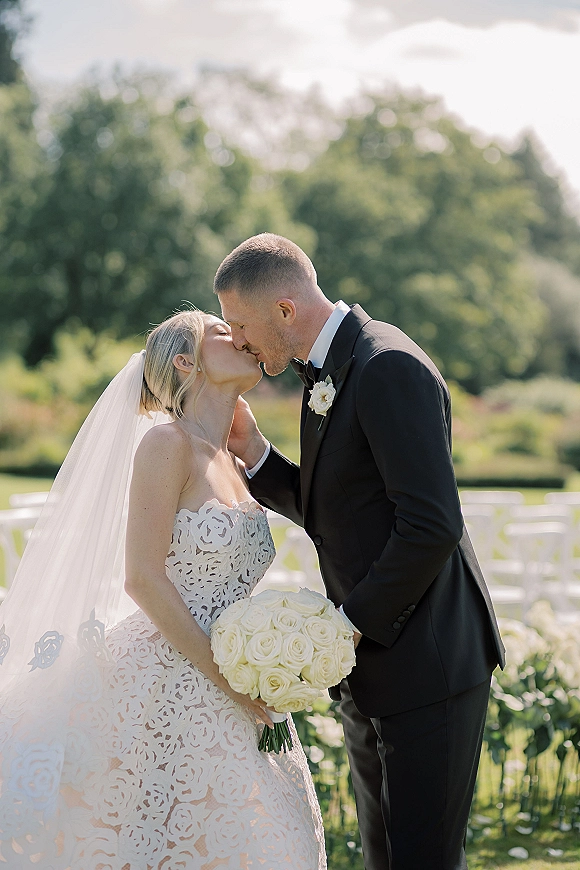 Wedding kiss portrait of bride and groom kissing, her veil and white rose bouquet visible, with garden trees and ceremony chairs behind them