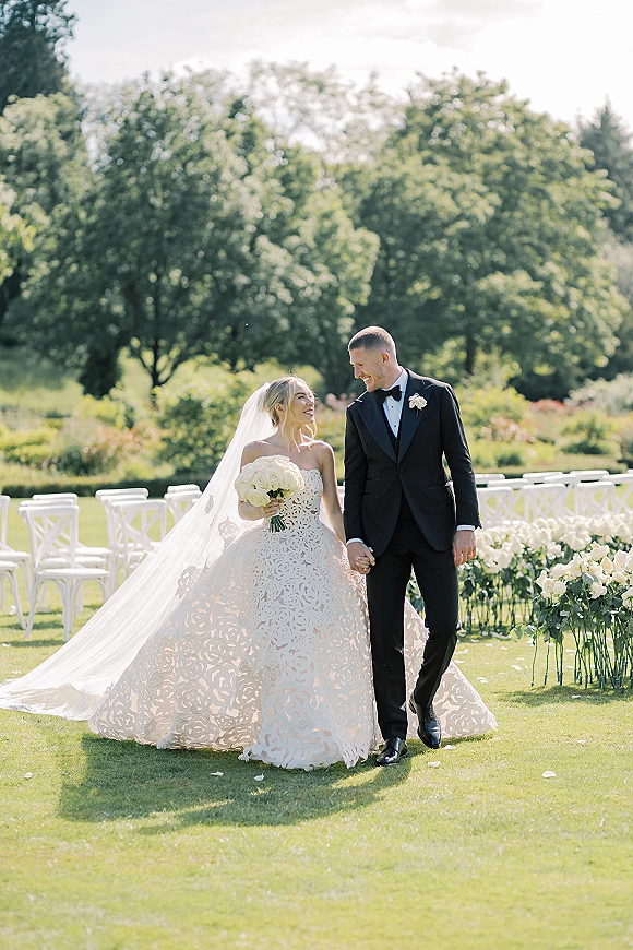 Couple portrait of bride and groom walking hand in hand, bride in strapless dress with long veil and white bouquet on a garden lawn aisle