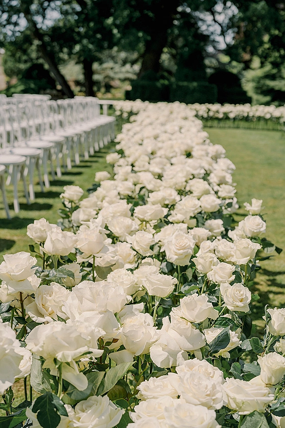 Ceremony aisle florals with white rose aisle arrangements lining a grass lawn path between white chairs, framed by garden trees and hedges
