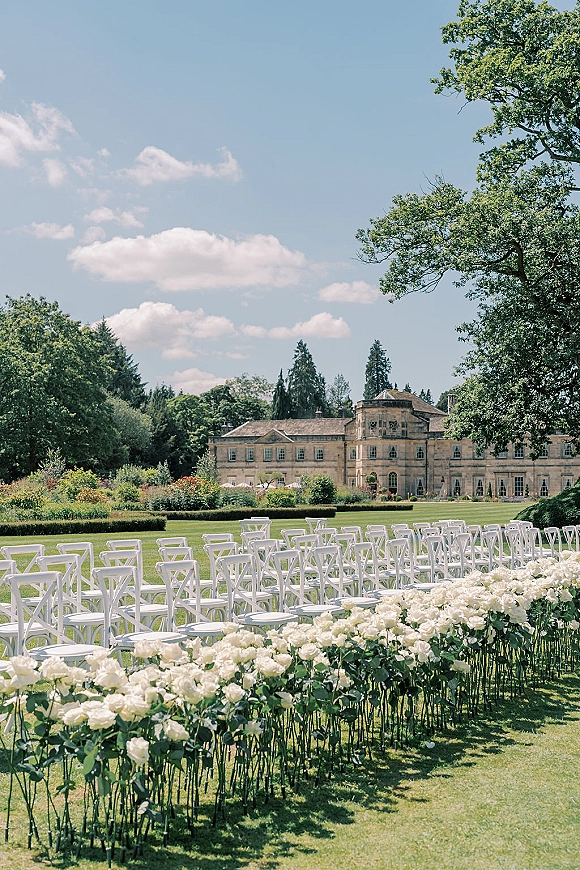 Outdoor ceremony setup with garden wedding ceremony rows of white folding chairs and white rose aisle flowers on a lawn by a manor house under blue sky