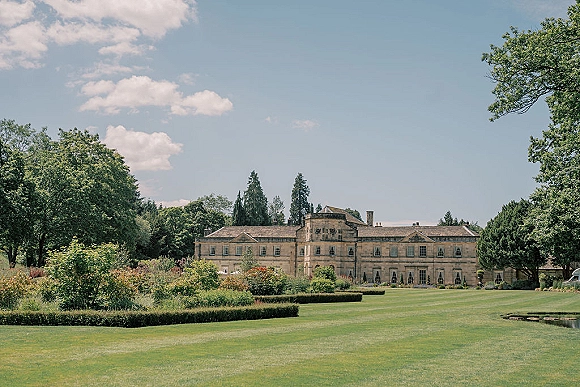 Wedding venue exterior of a manor house wedding venue with stone facade, trimmed hedges and manicured lawn beside a pond under blue skies