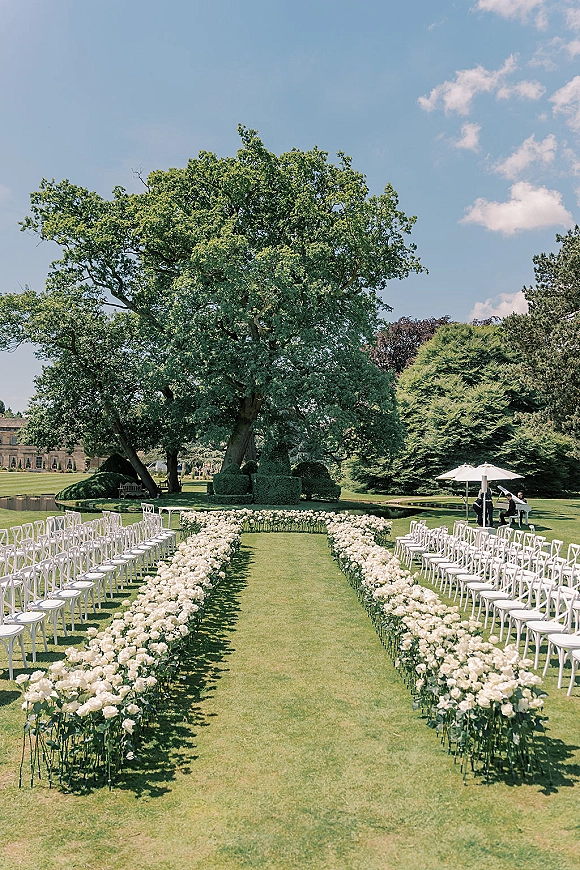 Outdoor ceremony setup with a garden wedding ceremony aisle lined in white flowers, rows of white chairs on an estate lawn under trees