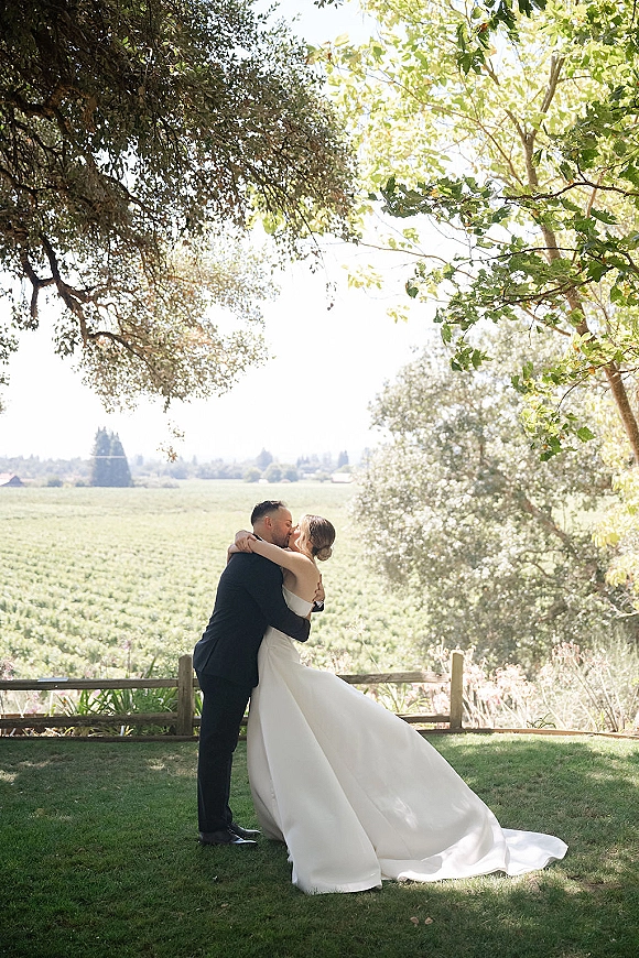 Wedding kiss portrait of bride and groom kissing, her strapless dress with long train by a wooden fence in a sunlit vineyard backdrop