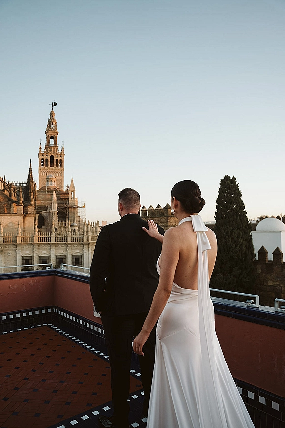 Wedding couple portrait from behind, bride and groom on a rooftop terrace with historic tower and skyline, her veil and bow dress detail visible