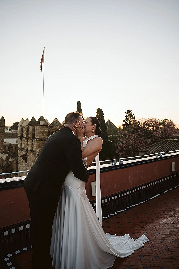 Wedding kiss portrait of bride and groom kissing as she holds his face, her gown train flowing on a rooftop terrace at sunset