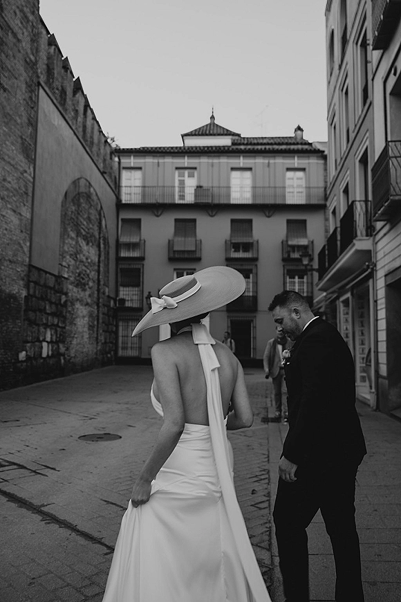 Couple portrait of bride in a wide brim hat and open-back dress with groom in black suit on a cobblestone city street