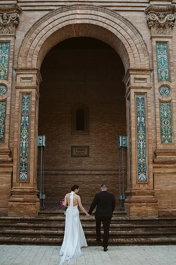 Couple portrait of bride and groom from behind holding hands, walking up stone steps under a brick archway, veil trailing