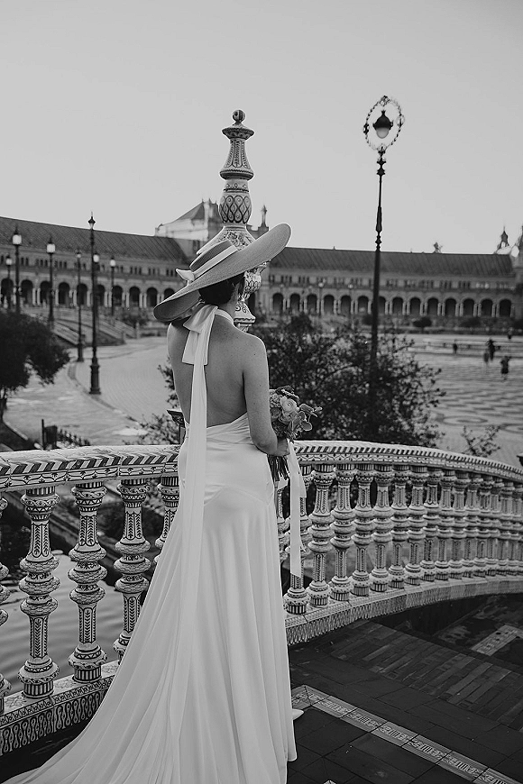 Bridal portrait of a bride from behind in a low-back wedding dress, holding a bouquet on an ornate tiled bridge with ceramic railing
