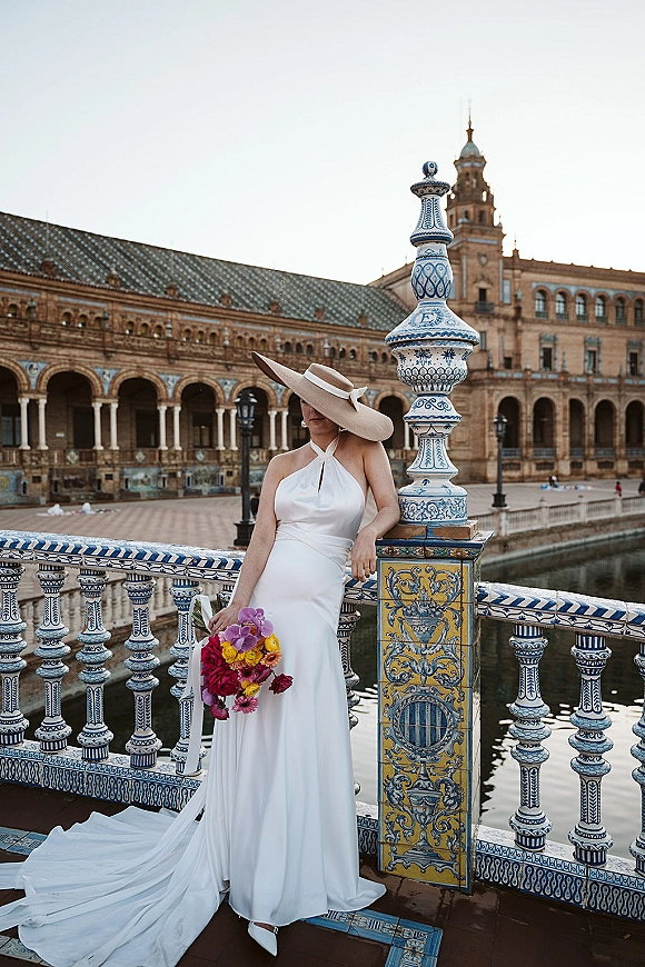 Bridal portrait of a bride in wide brim hat wearing a white satin dress with long train, holding a colorful bouquet on a blue tile bridge by a canal