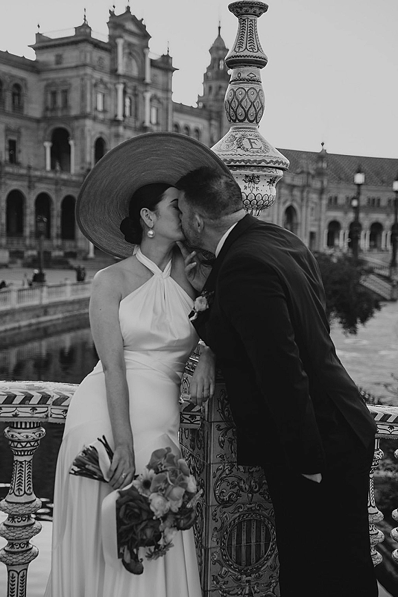 Wedding kiss in a black and white wedding photo as the bride in a wide brim hat and groom in dark suit lean on a tiled bridge railing by a riverfront historic building
