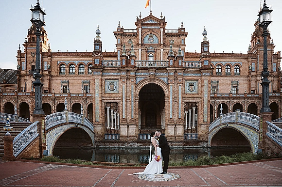 Wedding kiss portrait of bride and groom kissing, veil blowing as she holds a colorful bouquet by an arched canal bridge and ornate facade