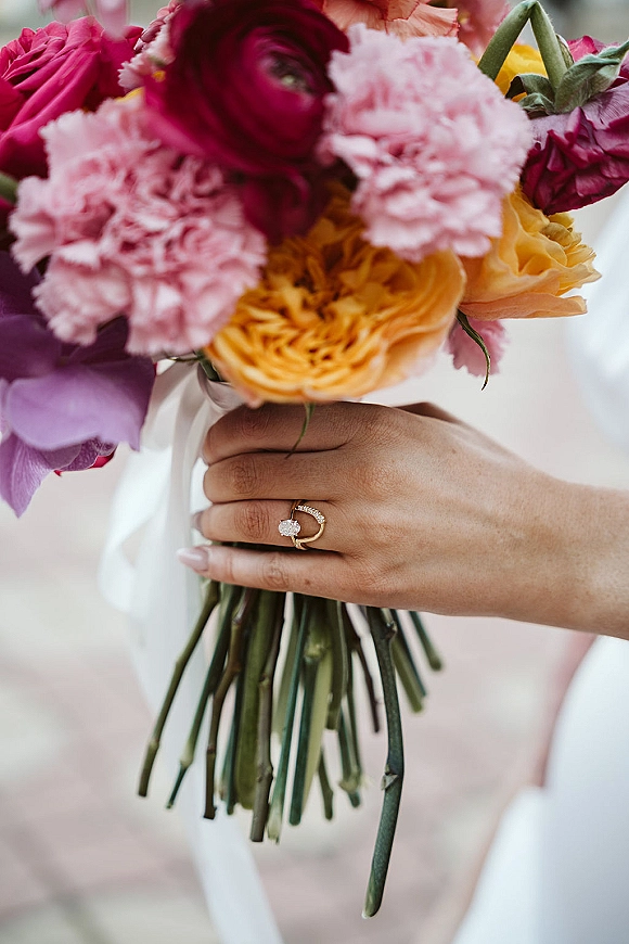 Bridal bouquet of bright wedding bouquet blooms in the bride’s hands, wrapped with a white ribbon, rings and manicured nails visible