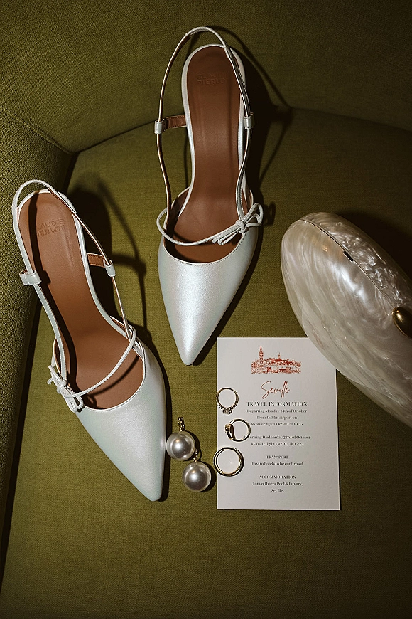 Bridal shoes styled as a flat lay with white slingback wedding heels, pearl drop earrings, rings and clutch on a green chair