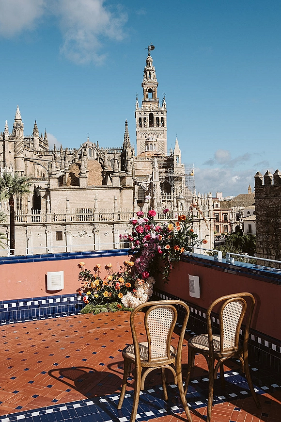 Rooftop wedding decor with rooftop wedding ceremony floral arrangement beside wicker chairs on tiled terrace, cathedral and skyline beyond