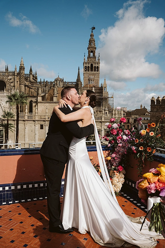 Wedding kiss portrait of bride and groom kissing, her long veil flowing as she holds a colorful bouquet on a rooftop terrace with cathedral towers behind