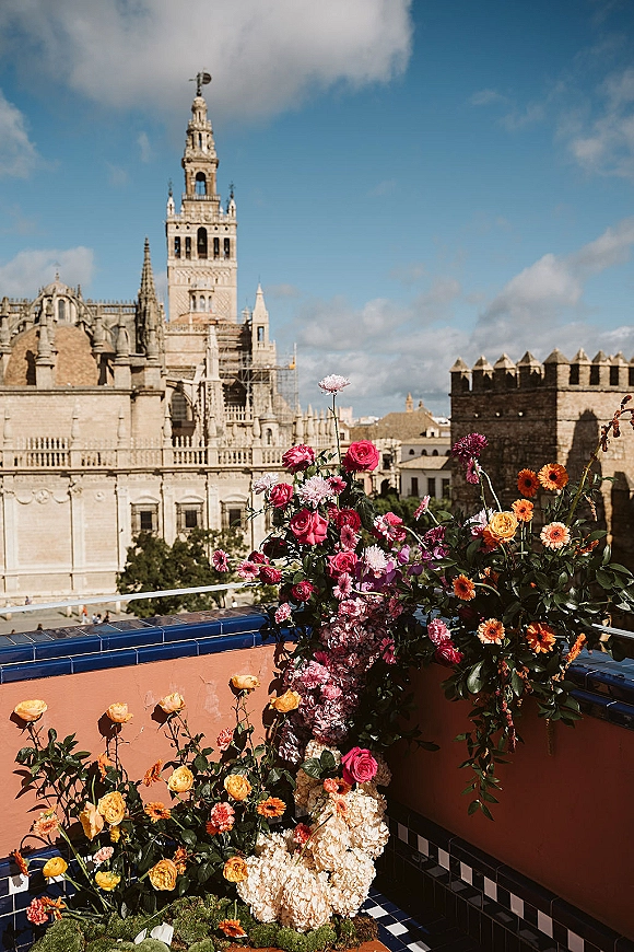Wedding floral installation with a ceremony flower arch of roses, hydrangea and gerbera daisies on a moss base by a cathedral terrace wall