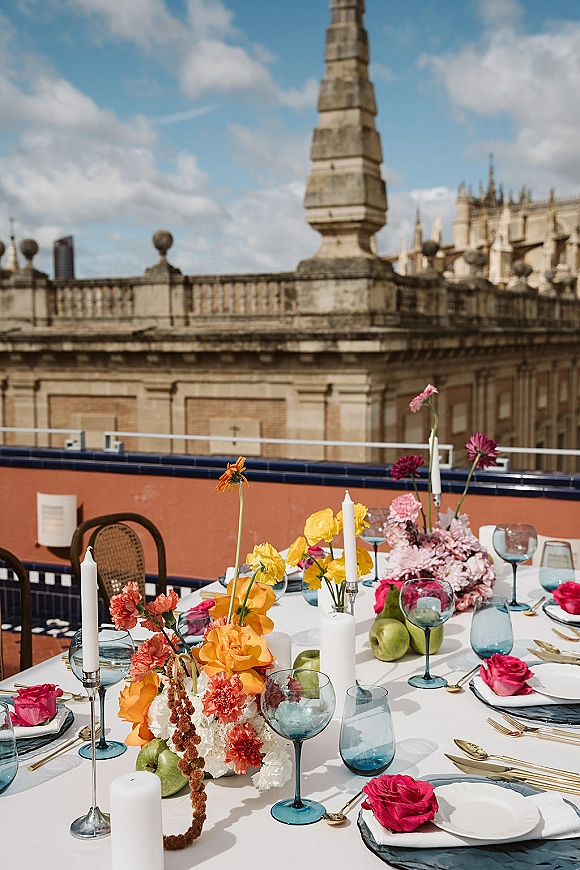 Reception tablescape with a colorful wedding tablescape centerpiece, tapered candles, blue goblets, gold flatware, and pears on a rooftop terrace.