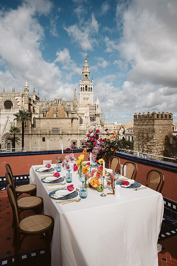 Reception tablescape for a rooftop wedding reception with white linen, colorful florals, blue goblets, and taper candles on a terrace skyline backdrop