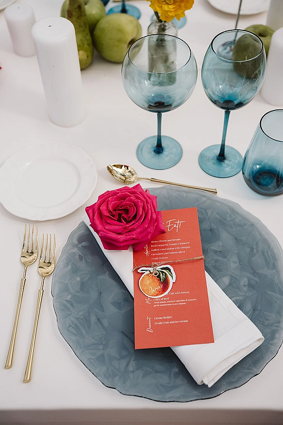 Reception tablescape with wedding place setting on a smoky blue glass charger, coral menu card, gold flatware, blue stemware, and candles