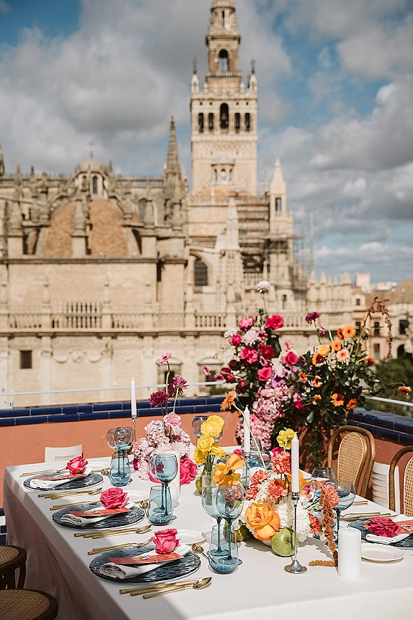 Reception tablescape with colorful wedding tablescape florals, taper candles, blue goblets, apples, and rattan chairs on a rooftop terrace skyline