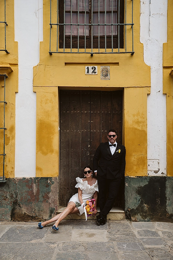 Couple portrait of bride seated in sunglasses holding a colorful bouquet beside groom in black tuxedo at a rustic wooden doorway