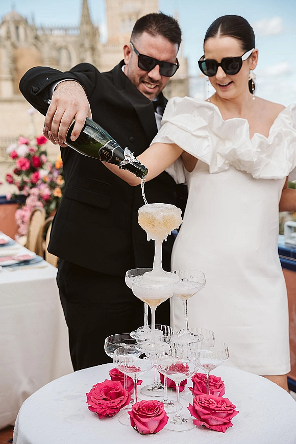 Champagne tower pour as bride and groom in sunglasses tip a bottle over coupe glasses with pink rose accent on an outdoor terrace