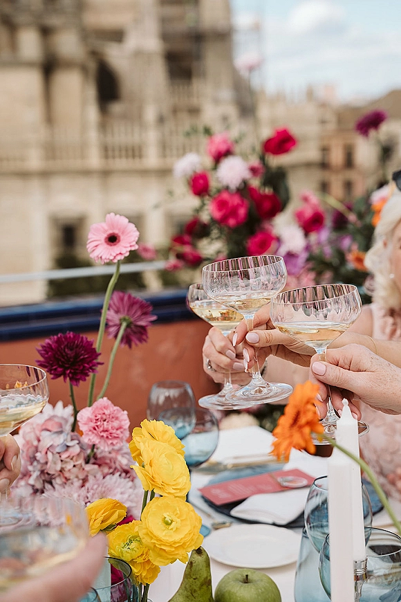 Wedding toast with champagne coupe glasses raised over a colorful reception tablescape on an outdoor terrace beside a stone facade