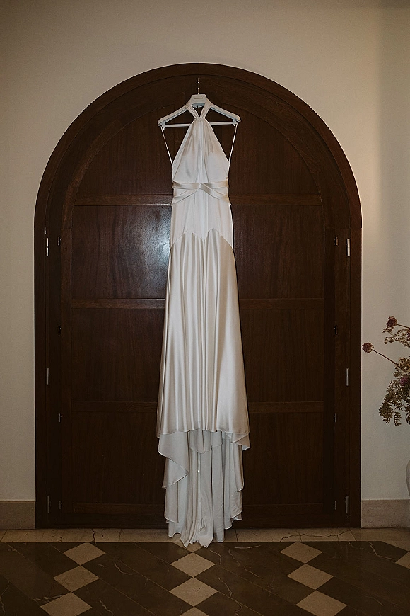 Wedding dress in satin wedding dress style hanging on a hanger before arched wooden doors, featuring a halter neckline and tiered skirt