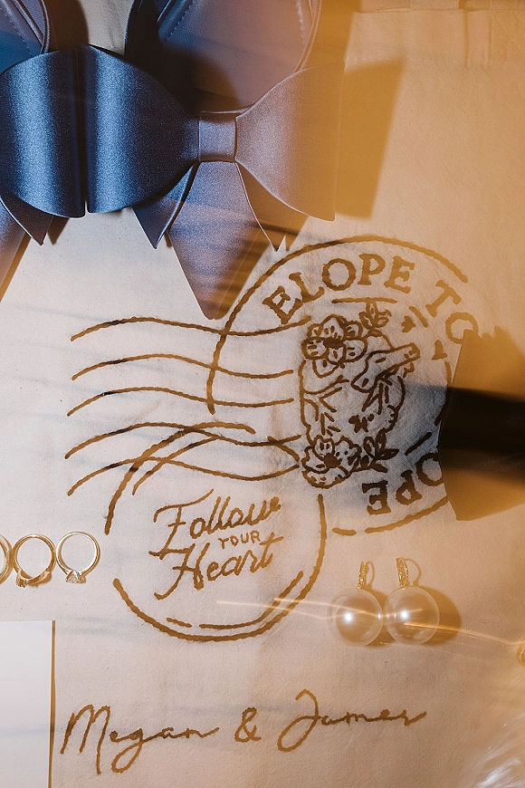 Invitation suite wedding invitation flat lay with navy bow tie, rings, pearl earrings, and postage stamp graphic on a white surface with warm light leaks