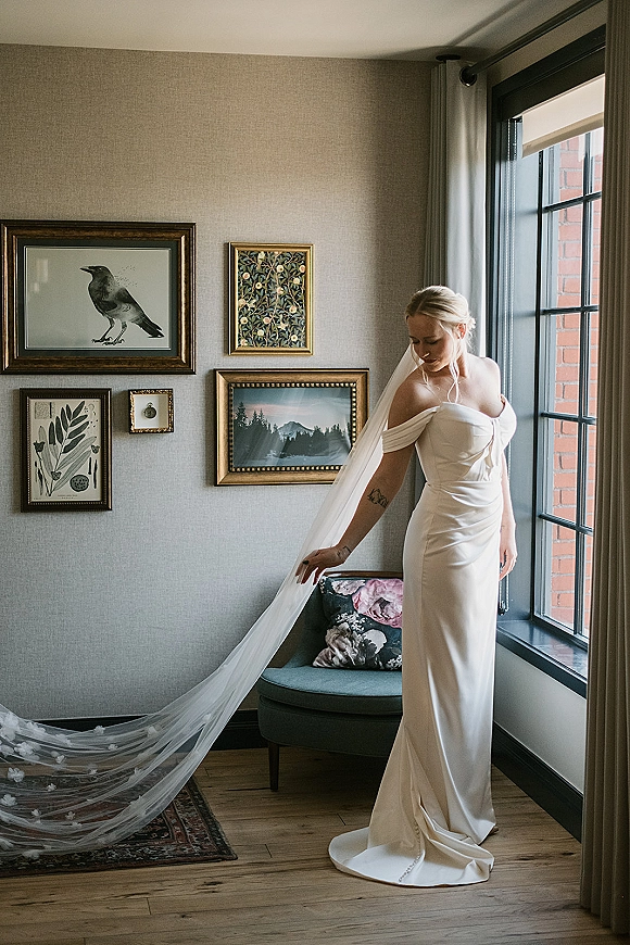 Bridal portrait of a bride in an off the shoulder wedding dress with long veil by a large window, framed art on gray wall behind