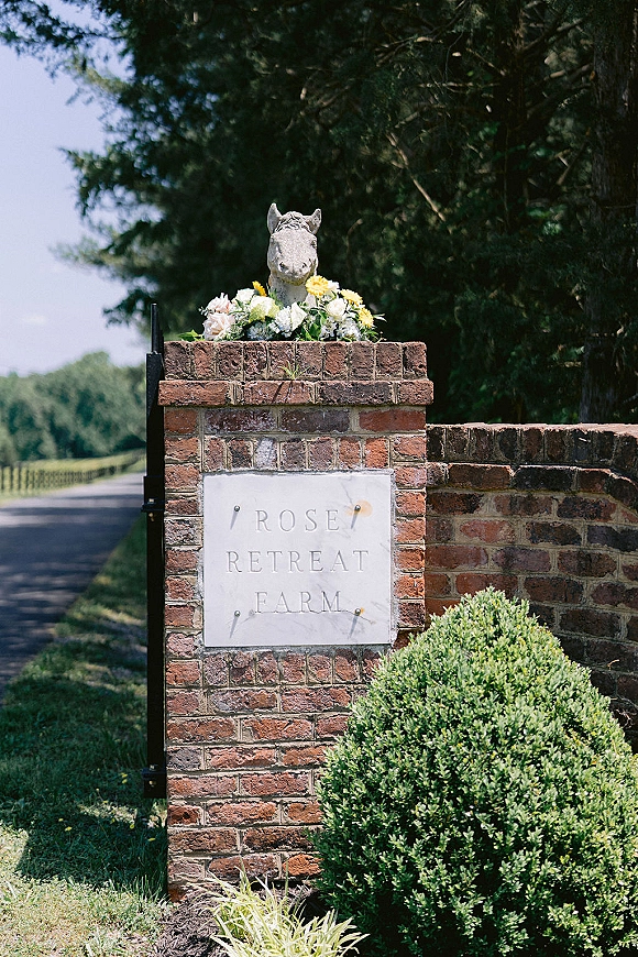 Wedding venue sign on a brick pillar with a floral arrangement atop a stone plaque, horse statue accent, and greenery by a country road
