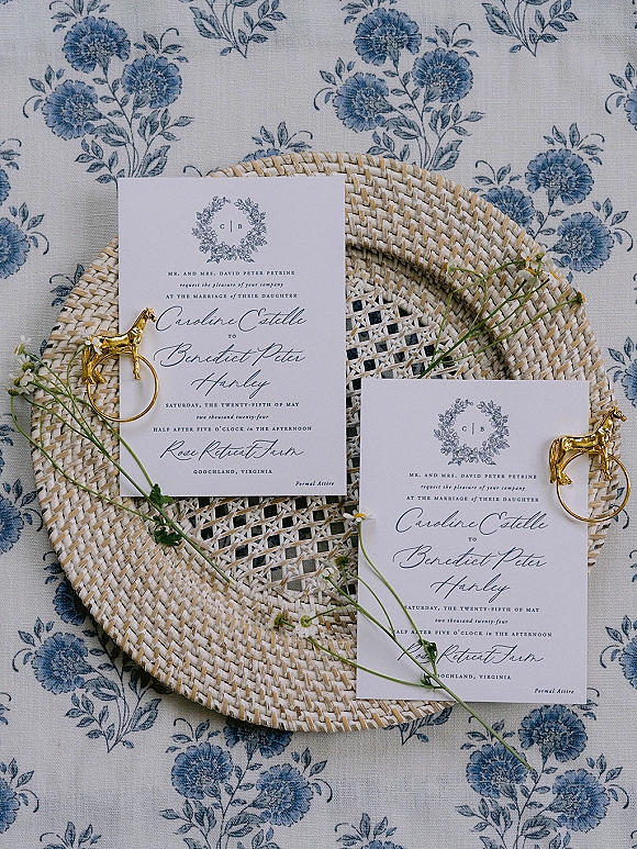 Invitation suite wedding invitation flat lay with calligraphy and monogram crest, gold horse napkin rings, wildflowers on blue floral tablecloth