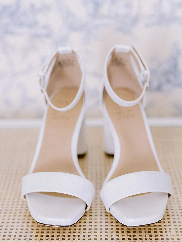Bridal shoes, white wedding heels with ankle strap buckles and open toes resting on a woven cane chair seat against a light wall