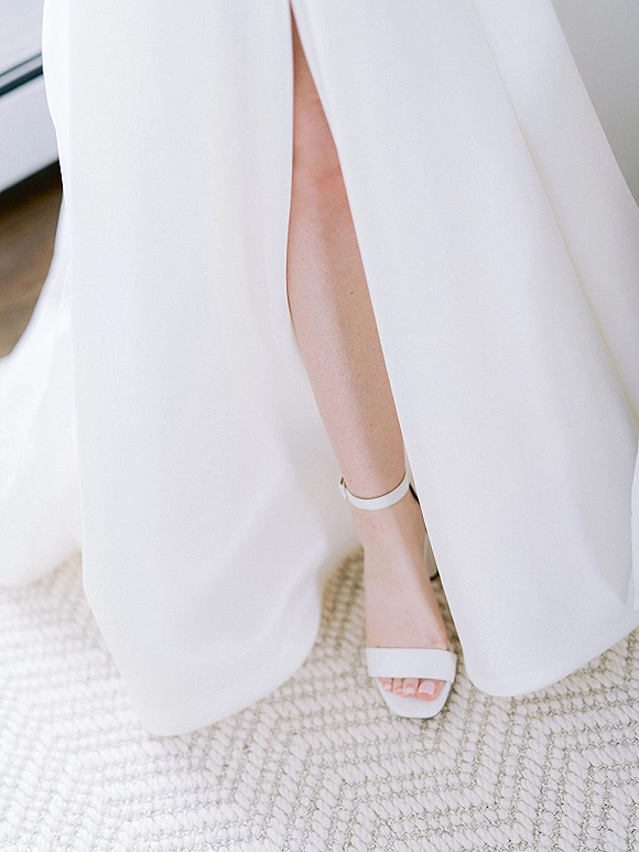 Bridal shoes, white wedding heels with ankle strap beside a bridal gown slit, resting on a woven rug indoors