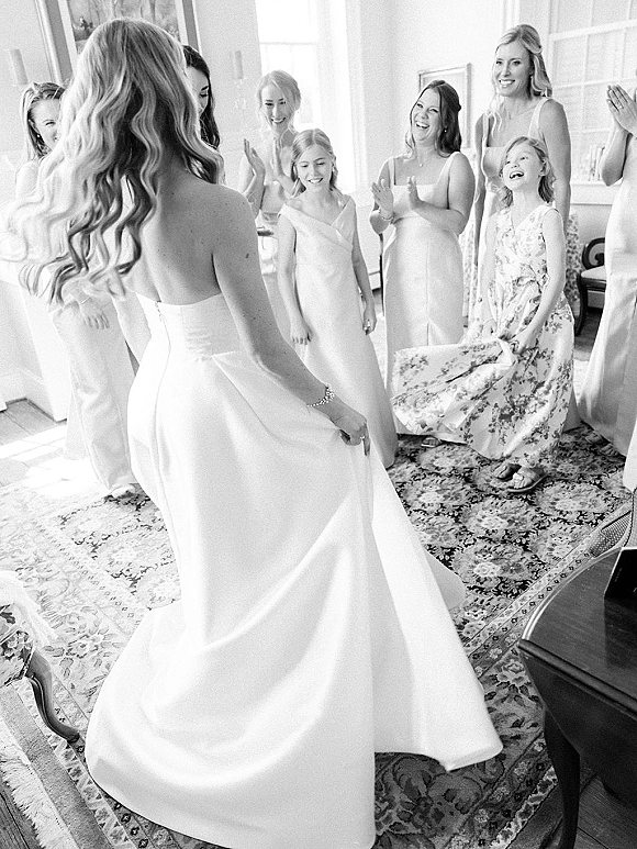 Getting ready moment as the bride reveal to bridesmaids in a strapless wedding dress, standing in a bright room with tall windows and piano