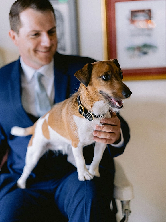 Groom with dog in a navy suit and tie, holding a small collared pup while seated indoors near a chair and framed artwork