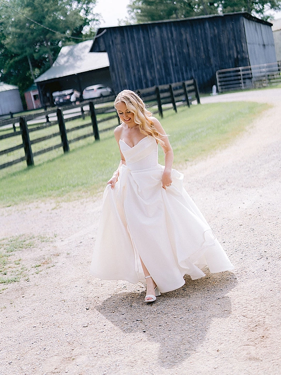 Bridal portrait of a bride holding her strapless satin gown with a high slit as she walks a gravel road past a barn and fence