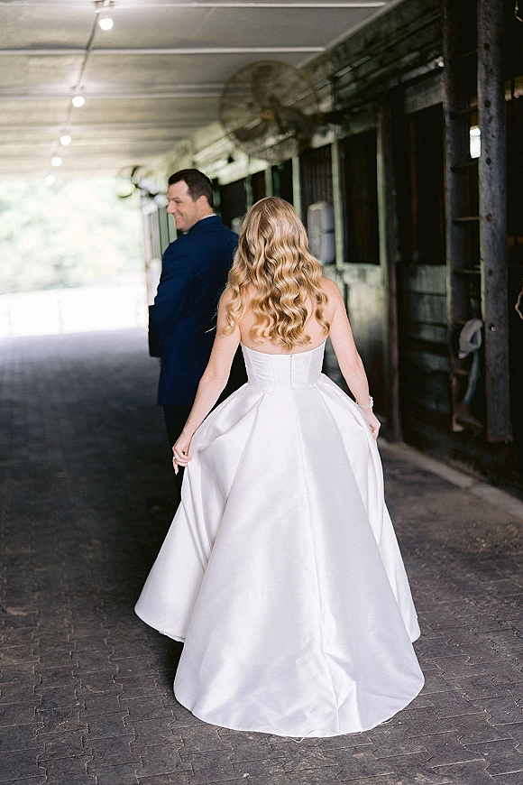 First look moment as bride in strapless satin ball gown walks up stable aisle toward groom waiting by barn stall doors under lights and fans