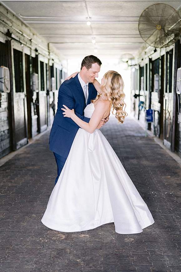 Couple portrait of bride and groom embrace in a stable aisle, her strapless satin ball gown and his blue suit under ceiling lights