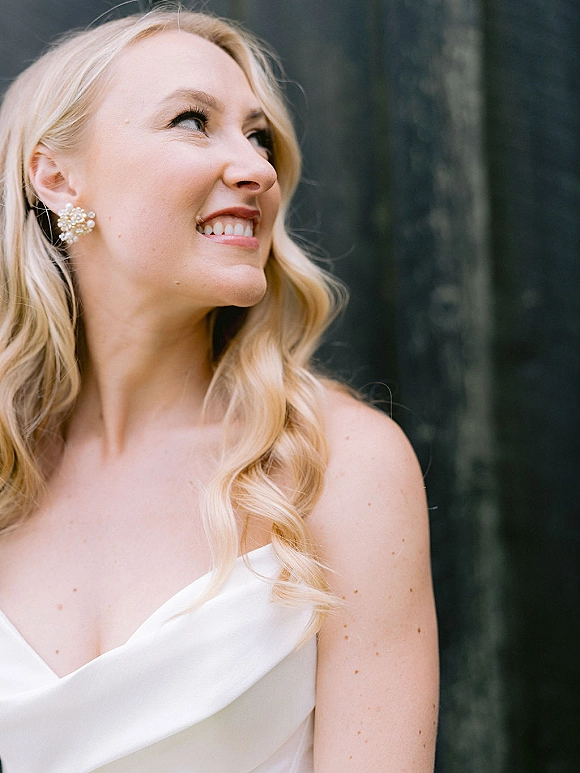 Bridal portrait of a smiling bride looking away in a strapless wedding dress with pearl cluster earrings against a dark wall