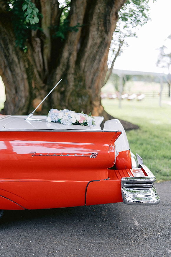 Wedding getaway car with blue-and-white floral arrangement on the hood, parked on a driveway near a large tree and outdoor pavilion