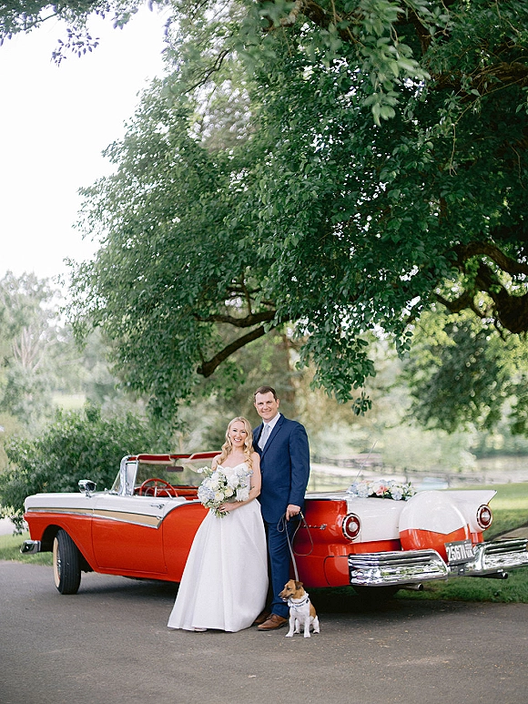 Couple portrait of bride and groom with car, bride in strapless gown holding white bouquet beside red vintage convertible under tree canopy