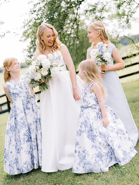 Bride with flower girls in blue and white dresses holding bouquets on a grassy lawn with trees and a wooden fence behind them