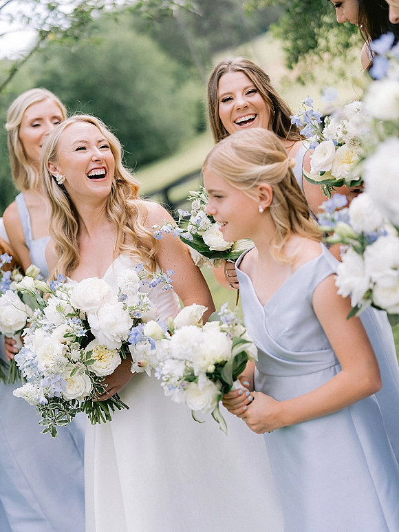 Bridesmaid group photo with bride and bridesmaids laughing, holding white and blue bouquets on a lawn with trees behind them