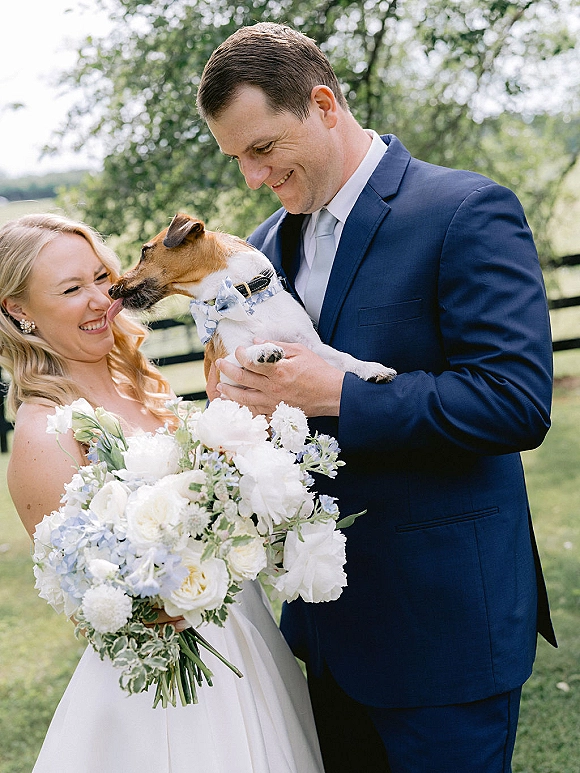 Couple portrait with bride and groom with dog as the pup licks the laughing bride, her bouquet held against a strapless dress on a grassy lawn by a wooden fence