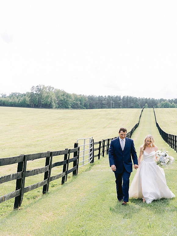 Couple portrait of bride and groom walking through a pasture field by a wooden fence, bride holding bouquet with a long veil trailing