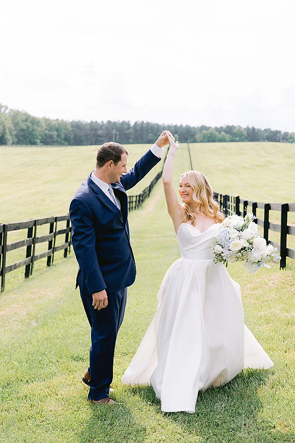 Couple portrait of bride and groom twirl while holding hands, her bouquet and strapless dress flowing in a field by a rustic fence