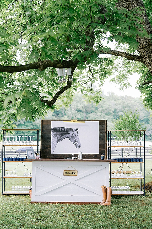 Wedding bar setup with outdoor wedding bar shelves, glassware and personalized bar sign, framed horse art and boots under lanterns on lawn